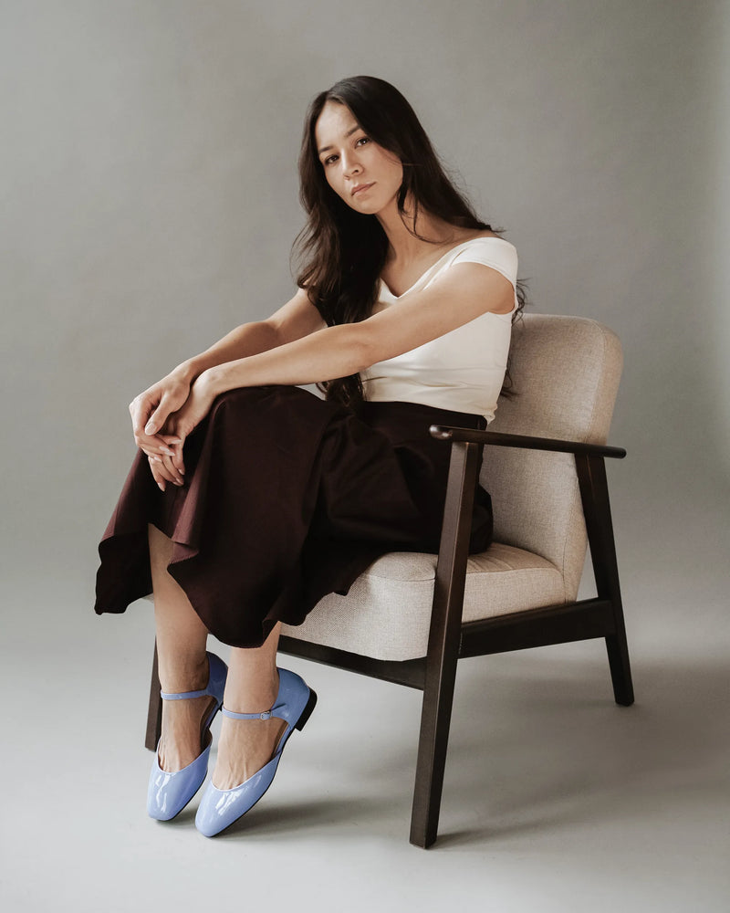 Full-body shot of a seated model wearing the flats with a skirt. The Gallery Mary Jane Cornflower Patent. Sizes 5–12.