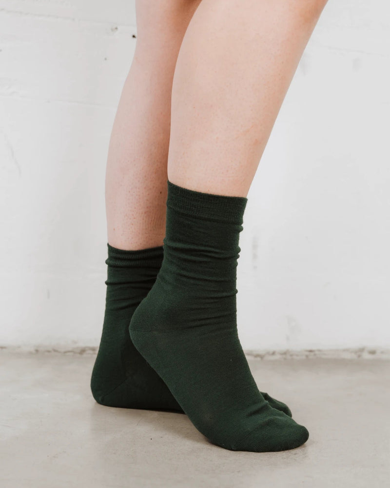 Side angle, feet crossed, wool knit with a folded ribbed cuff. The Dressy Crew Sock Balsam.
