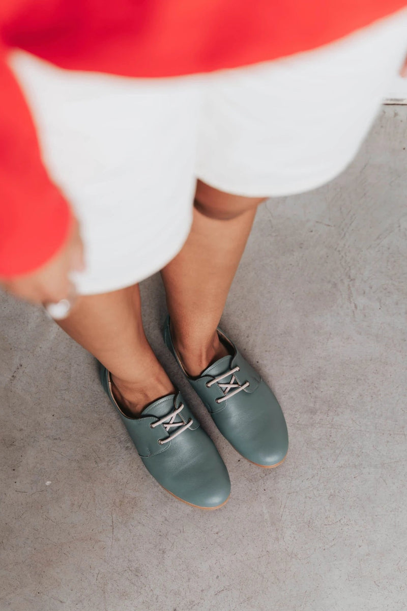 Feet shown wearing lace-up oxford flats. The Eyelet Oxford 2.0 Storm. Sizes 5–12, wide available.