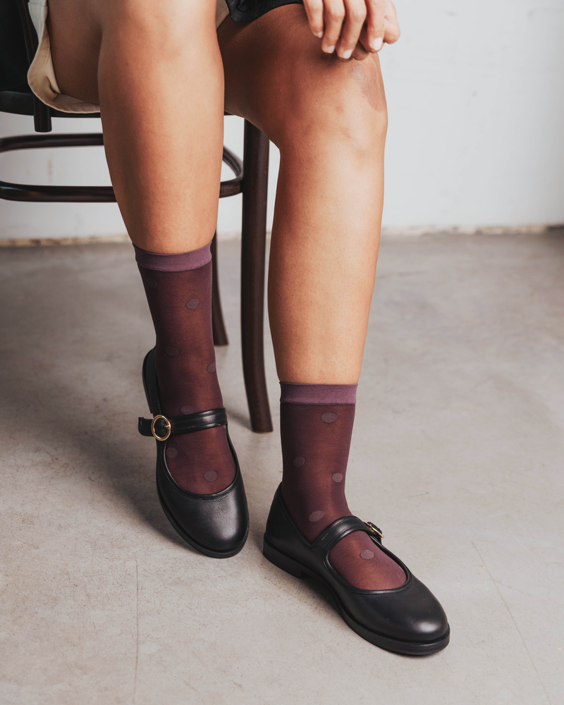 Seated close-up of legs wearing sheer dotted socks with mary jane flats. Rachel Mega Dot Sheer Socks in Plum.