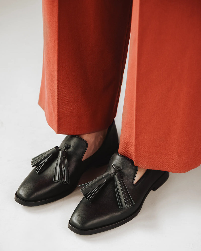 Woman wears leather tassel loafers with rust trousers. The Tassel Daily Loafer Black. Sizes 5–12, available in wide.
