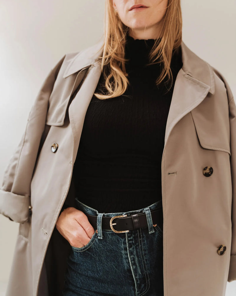 Black leather belt with gold buckle, styled with jeans, black turtleneck, and beige trench coat. The Accent Belt Gold Black.