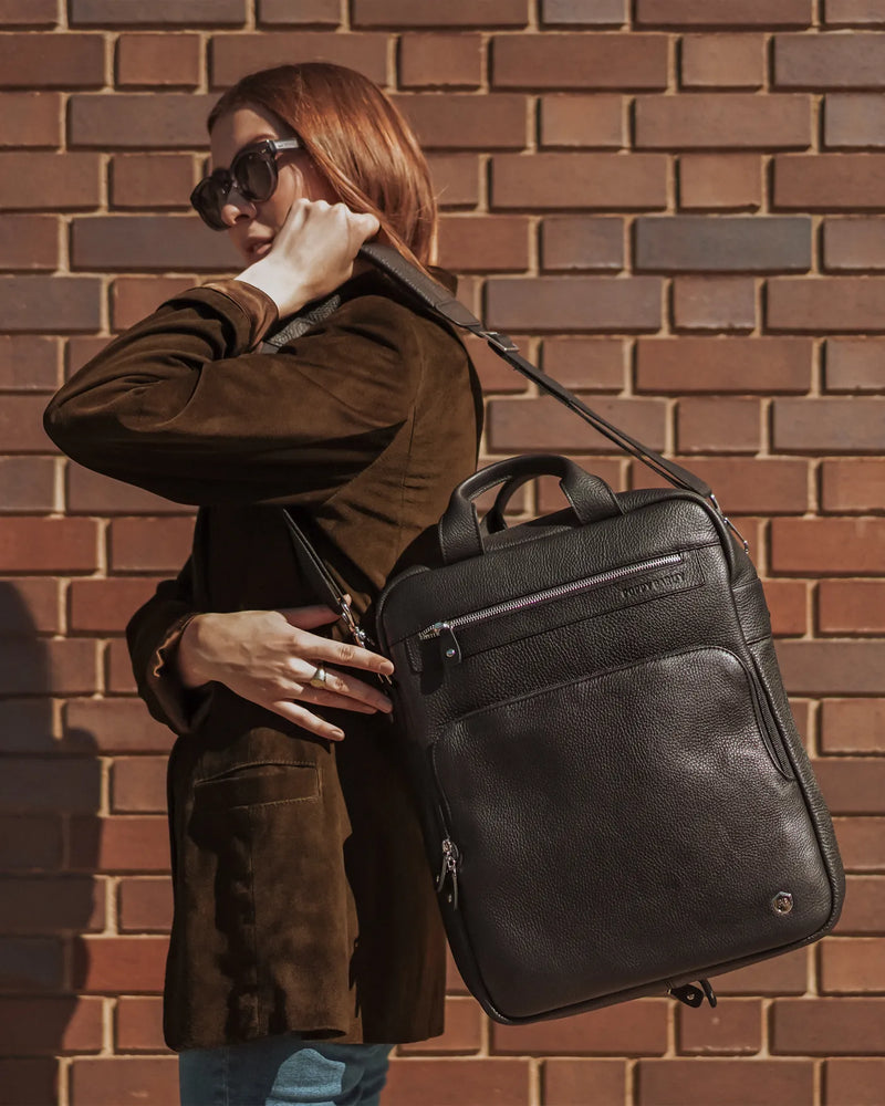 The Backpack Black in pebbled leather worn crossbody by woman in brown suede jacket against brick wall