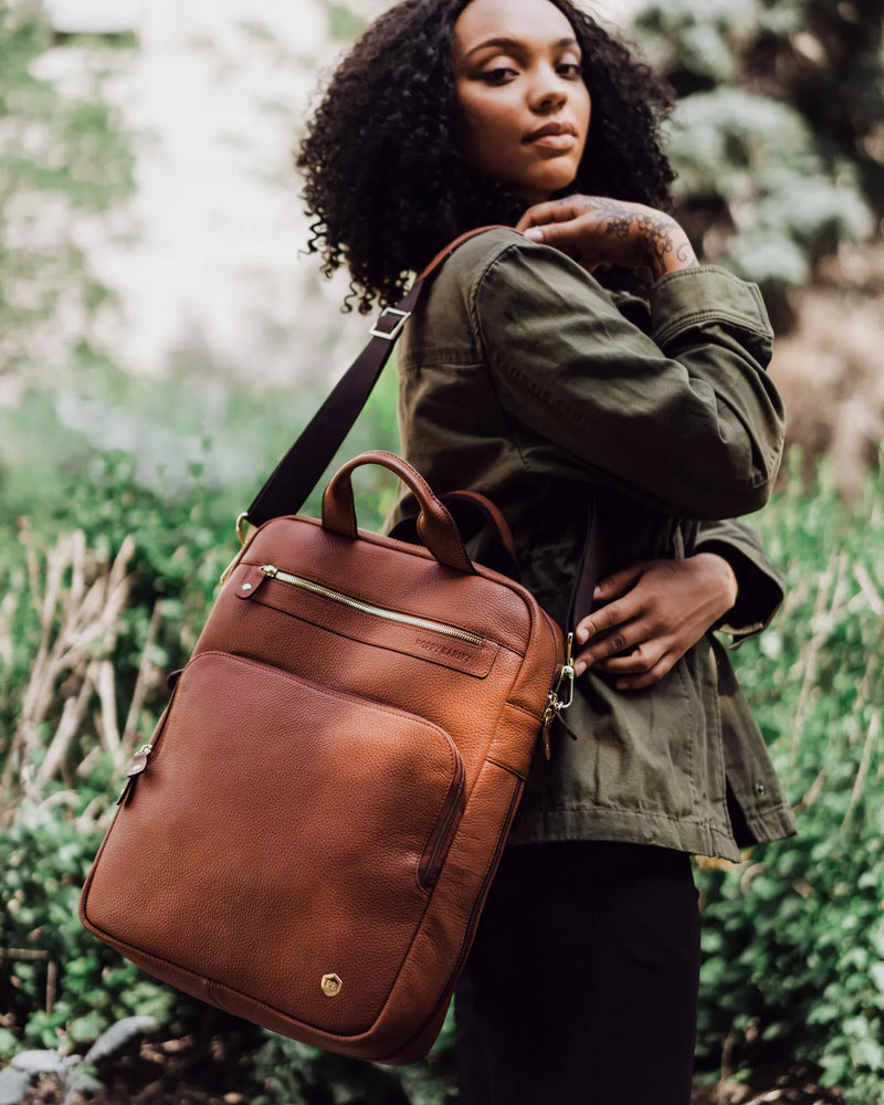 Woman in olive jacket carrying pebbled leather backpack with gold hardware outdoors. The Backpack Hazelnut.
