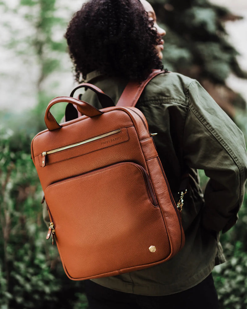 Woman wearing pebbled leather backpack with gold hardware, olive jacket, outdoors. The Backpack Hazelnut.