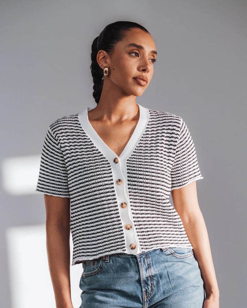 Woman wears a black stripe short-sleeve knit cardigan with V-neck and wooden buttons. The Beach House Cardigan Black Stripe.
