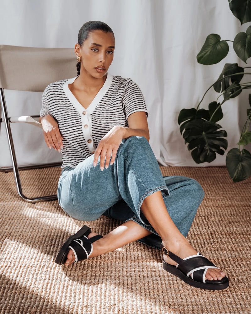 Woman seated on a rug in a black stripe knit cardigan, jeans, and black sandals. The Beach House Cardigan Black Stripe.