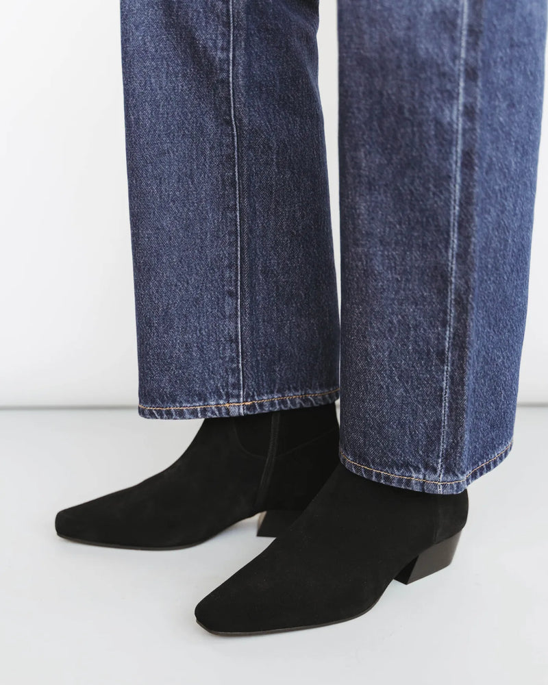 Woman in dark jeans wears suede square-toe ankle boots with low block heel. The Beau Boot Black. Sizes 5–12.