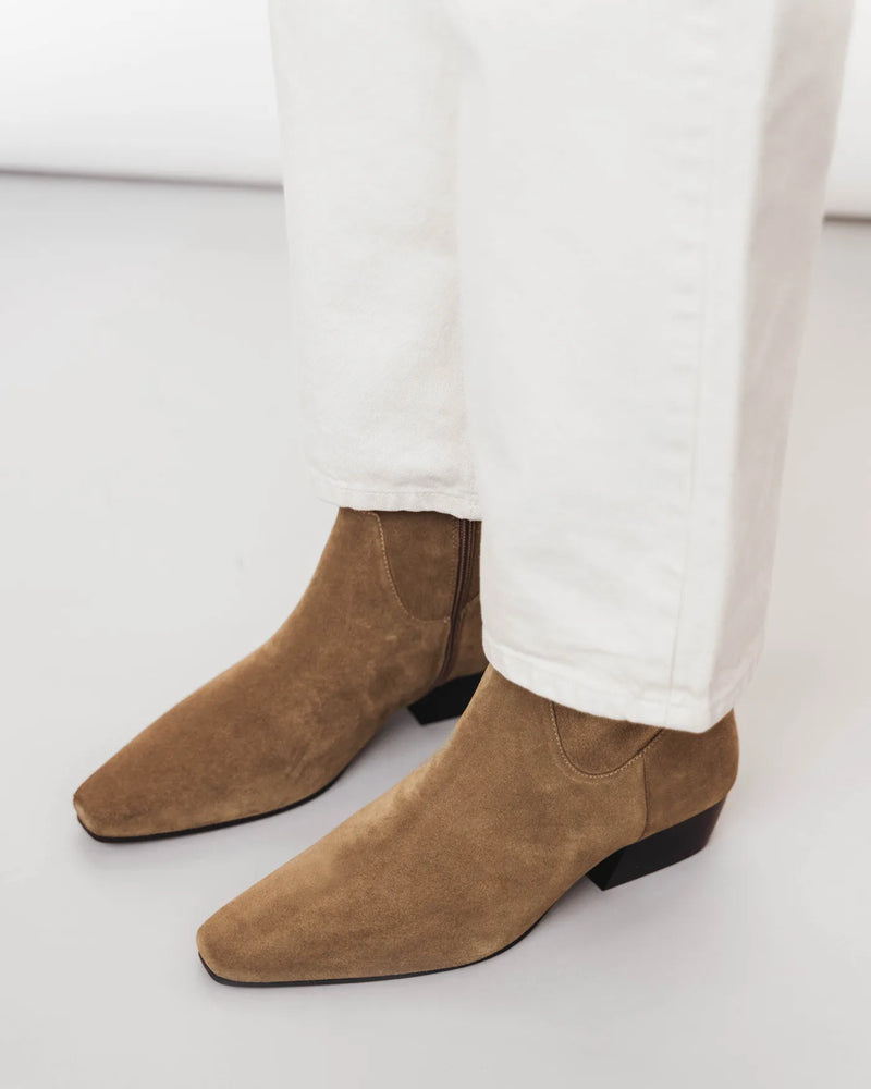 Woman in white jeans wears suede square-toe ankle boots with low block heel. The Beau Boot Desert Suede. Sizes 5–12.