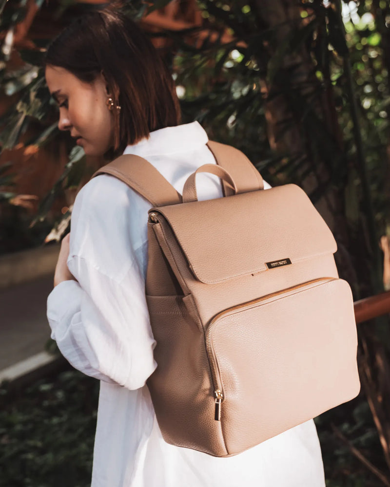 Woman carrying a pebbled leather backpack outdoors amid tropical greenery. The Beyond Backpack Biscotti.
