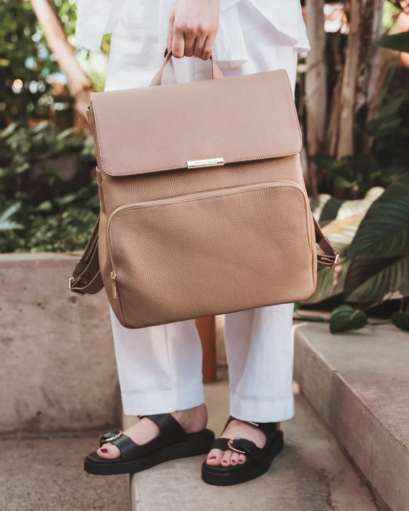 Woman holding a pebbled leather backpack by the top handle on outdoor steps. The Beyond Backpack Biscotti.