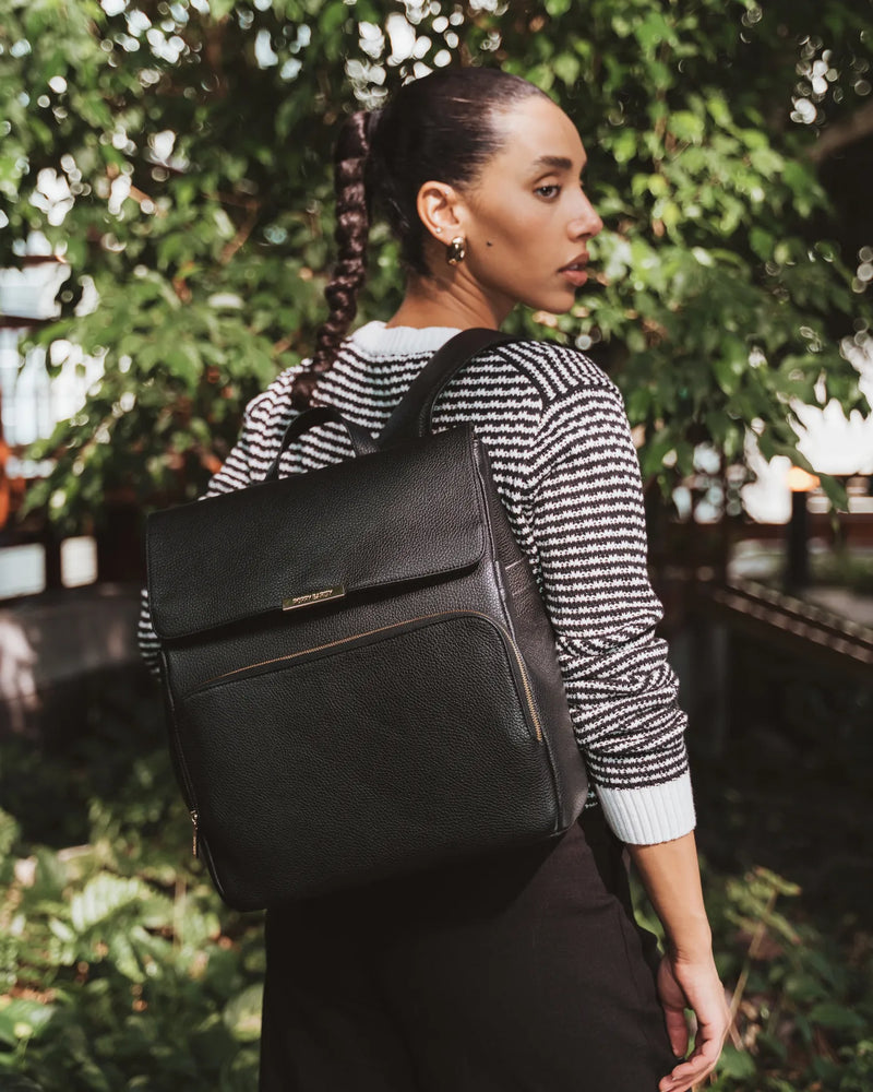 Woman in striped sweater wears a pebbled leather backpack outdoors among lush greenery. The Beyond Backpack Black.