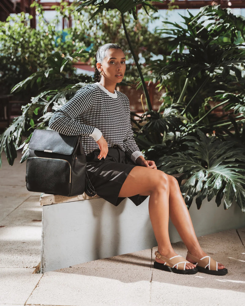 Woman seated outdoors in striped top and black skirt holding a leather backpack. The Beyond Backpack Black.