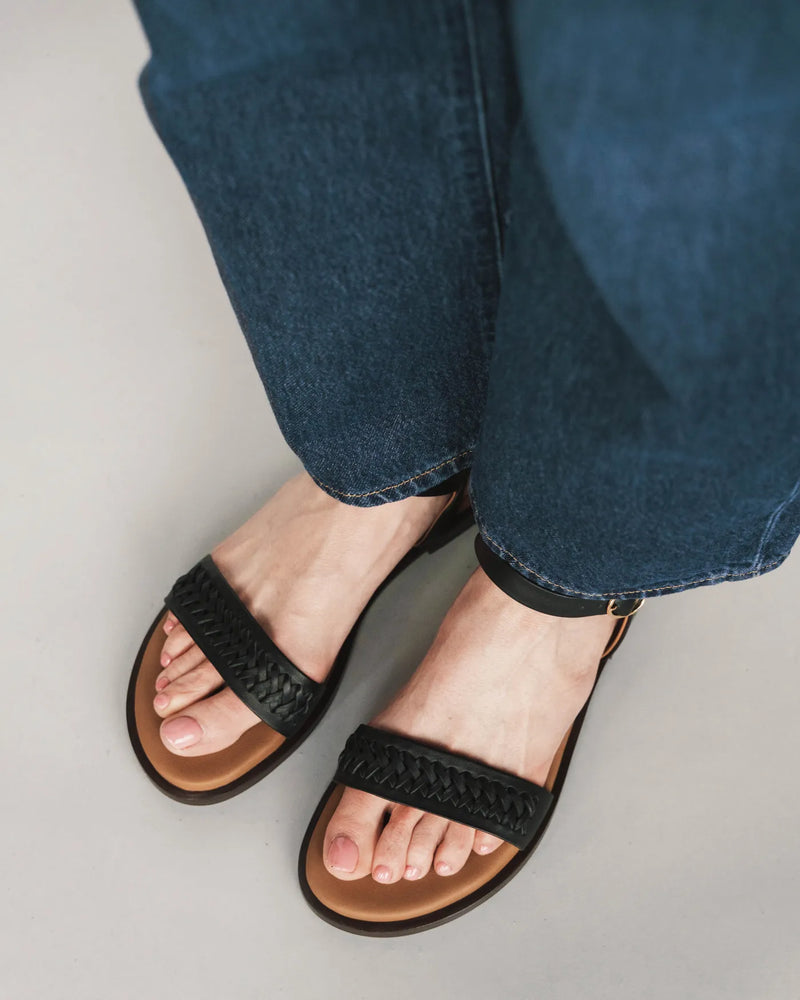 Woven leather flat sandals with ankle strap, worn with jeans. The Bohème Sandal Black. Sizes 5–12, available in wide.