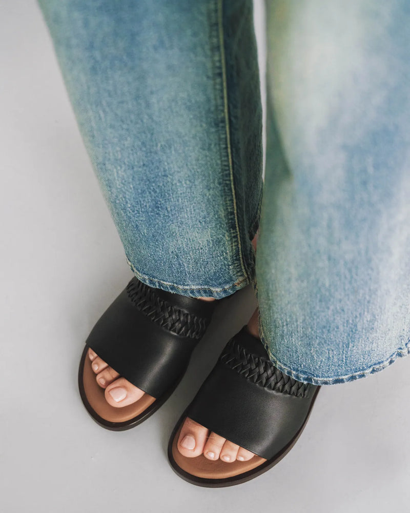 Leather slide sandals with woven toe strap detail, overhead view with light jeans. The Bohème Slide Black. Sizes 5–12.