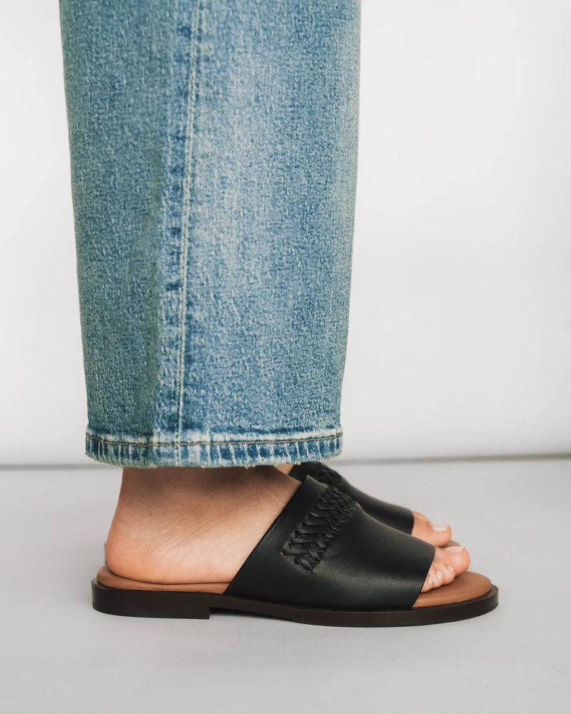 Leather slide sandals with woven toe strap, side view with light wash jeans. The Bohème Slide Black. Sizes 5–12.