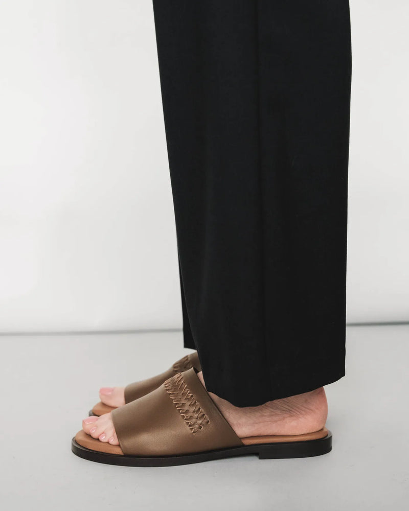 Leather slides with woven detail, side view, black pants. The Bohème Slide Tapenade. Sizes 5–12, available in wide.