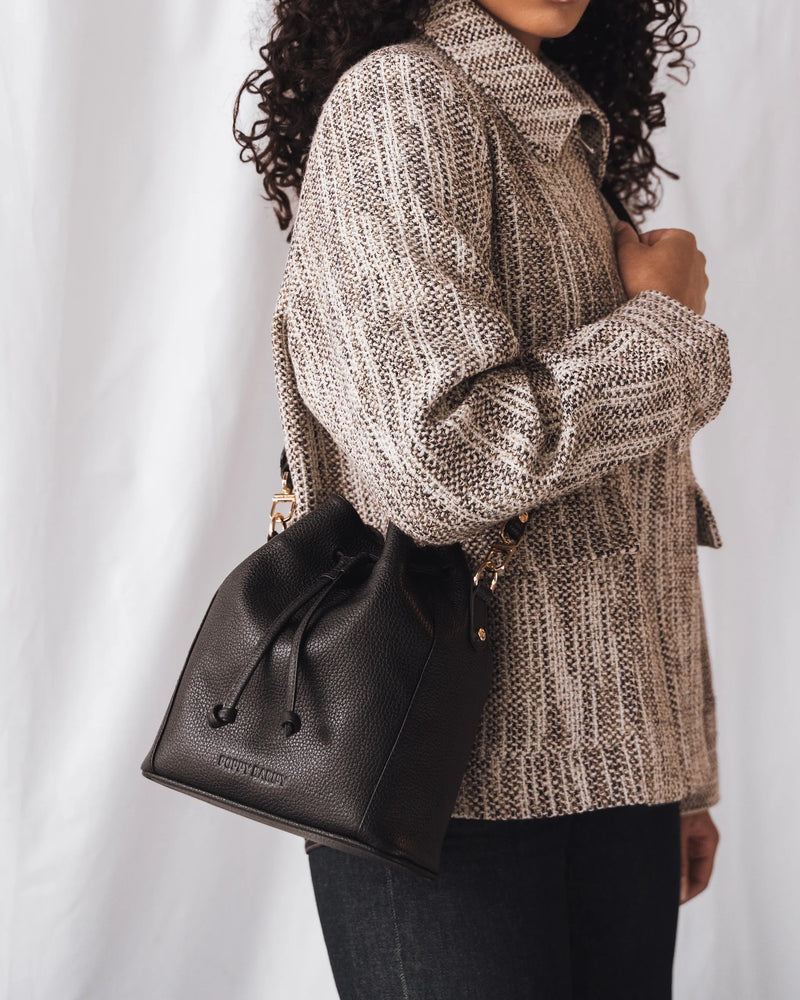 Woman in chunky knit sweater wears a pebbled leather drawstring bucket bag as a crossbody. The Bondi Bucket Bag Black.