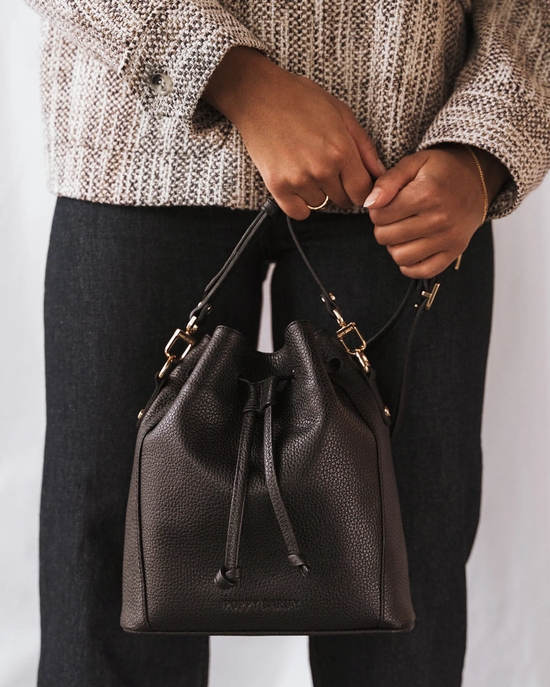 Woman in chunky knit sweater holds a pebbled leather drawstring bucket bag by its straps. The Bondi Bucket Bag Black.