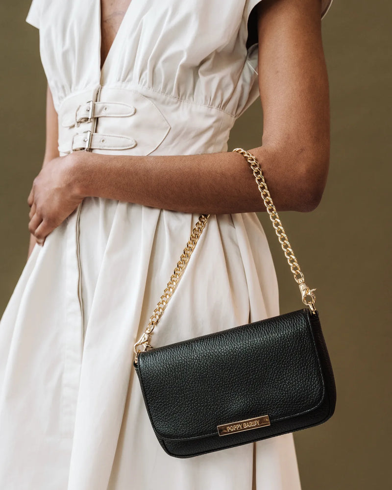 Woman in white belted dress carries small black pebbled leather flap bag on a gold chain strap. The Chain Strap.