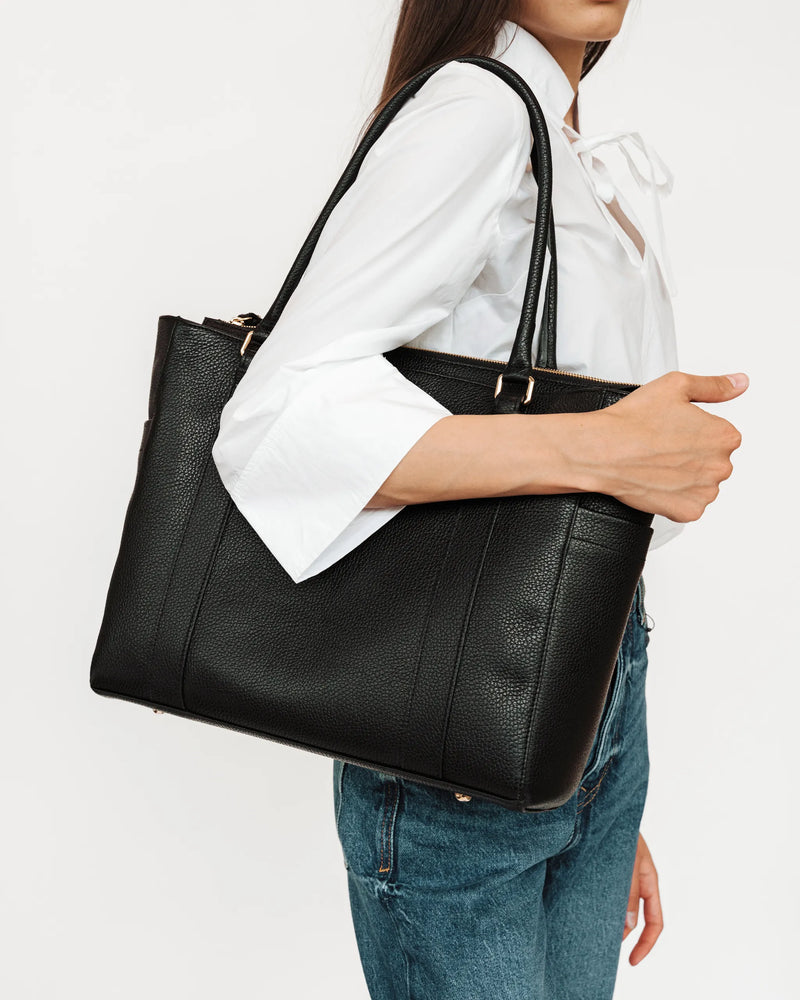 Woman in white tie-neck shirt and jeans carries large pebbled leather tote over shoulder. The Colleague Tote Black.