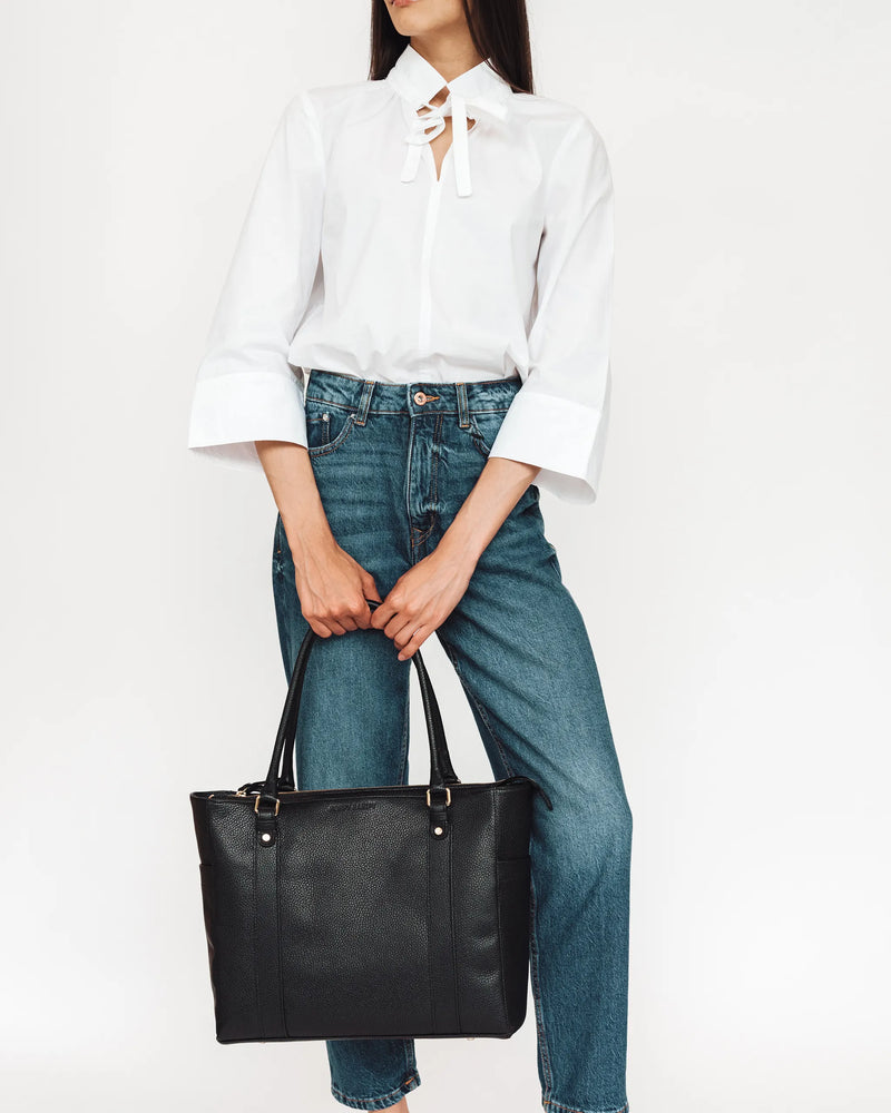 Woman in white shirt and jeans holds pebbled leather tote by handles at her side. The Colleague Tote Black.