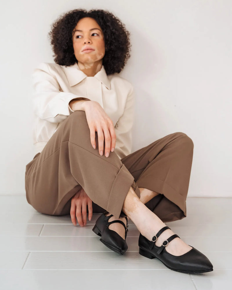 Double-strap Mary Jane flats on woman in cream jacket and taupe trousers. The Editorial Mary Jane Black. Sizes 5–12.