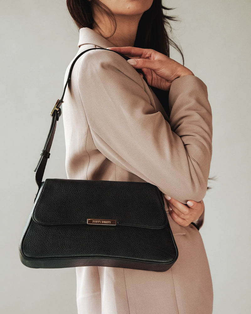 Woman in blush coat wears pebbled leather flap shoulder bag with gold logo. The High Line Shoulder Bag Black.