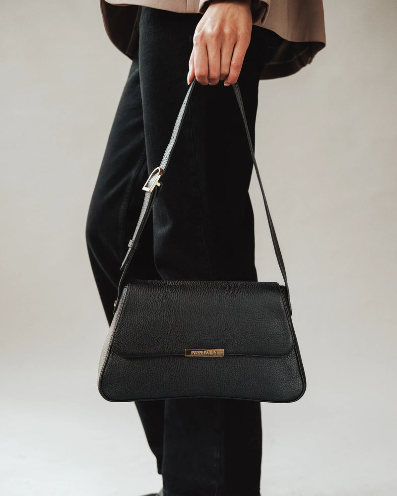 Woman pants holds pebbled leather flap shoulder bag by its single strap. The High Line Shoulder Bag Black.