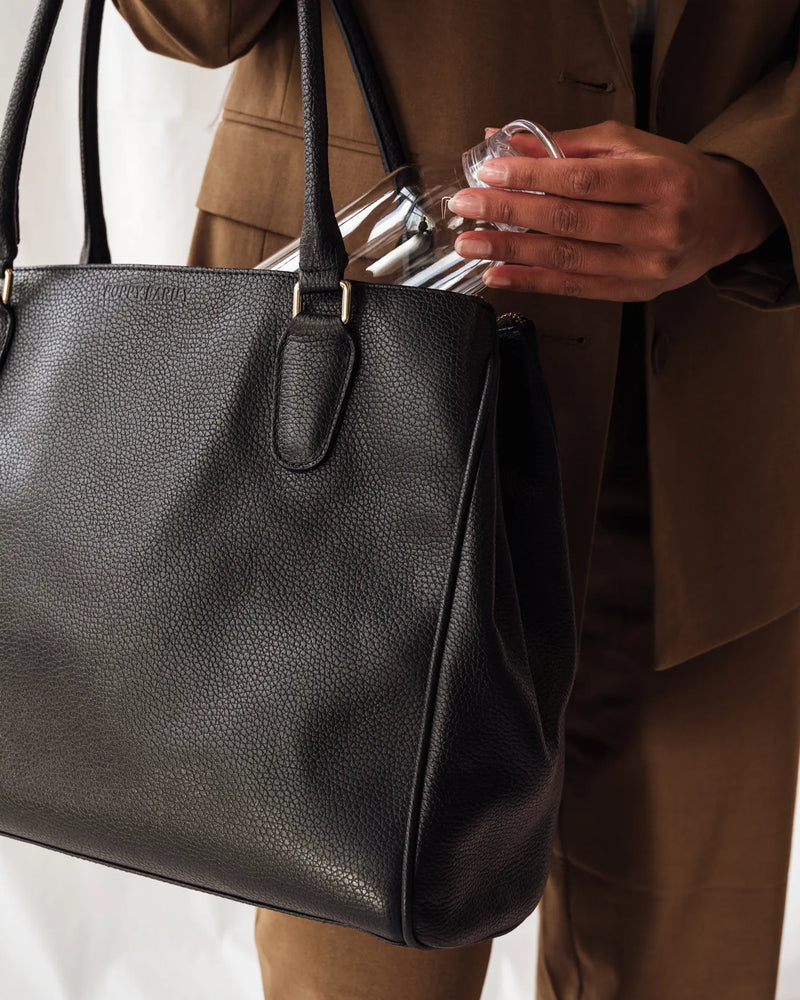 Woman in tan blazer placing a clear Kinto water bottle into a large black leather tote. The Kinto Water Bottle Clear.