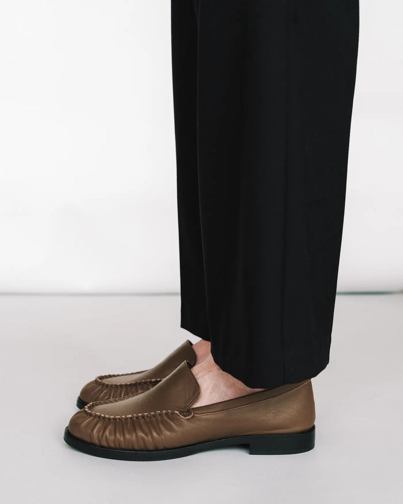 Leather loafers with ruched toe, seen from the side with black trousers. The Laid Back Loafer Tapenade. Sizes 5–12.