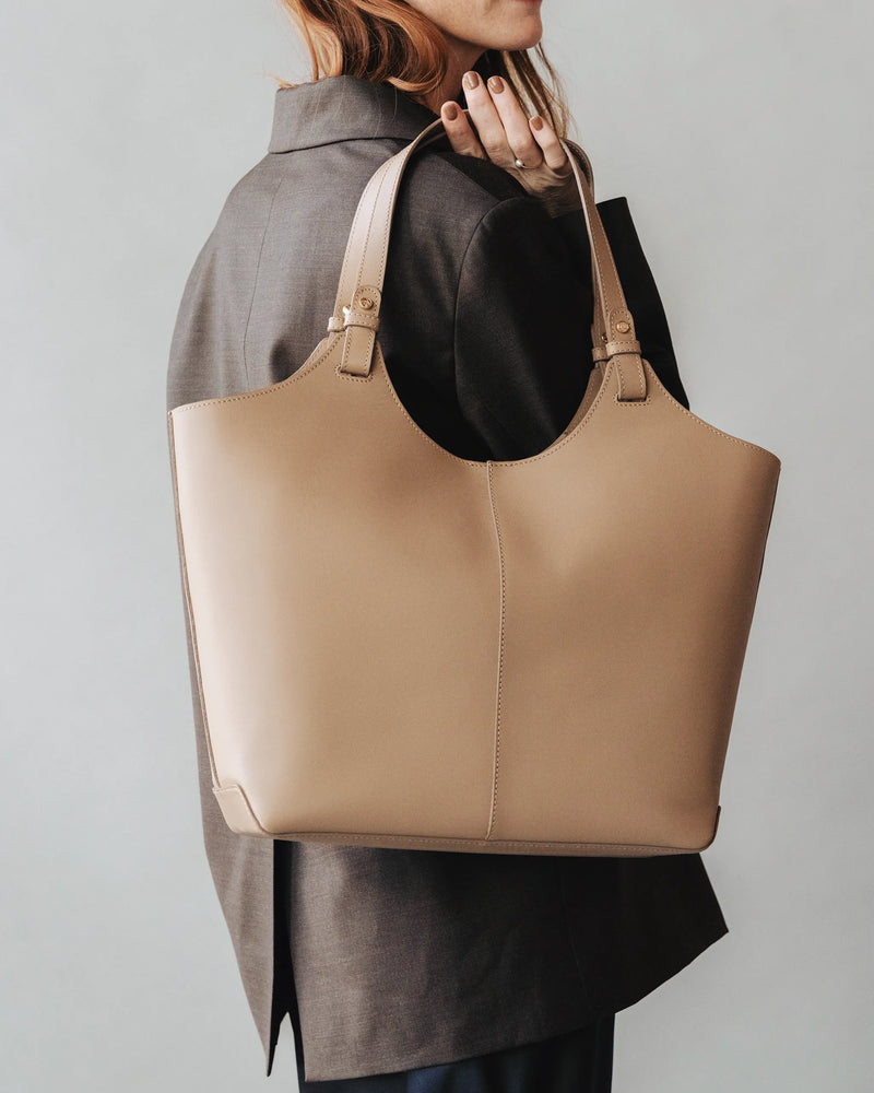 Woman in gray blazer carries a large leather tote on her shoulder. The Lisboa Tote Biscotti.
