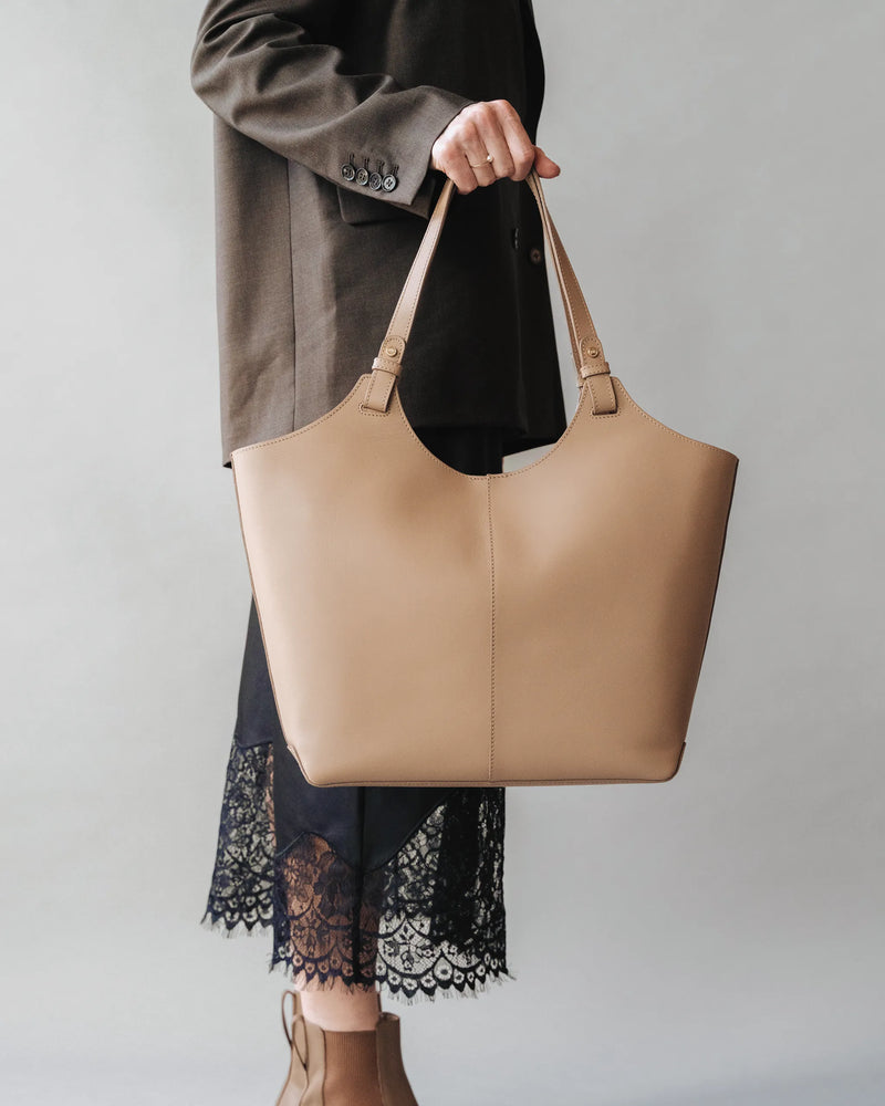 Woman in gray blazer and lace-hem dress carries a large leather tote by the handles. The Lisboa Tote Biscotti.