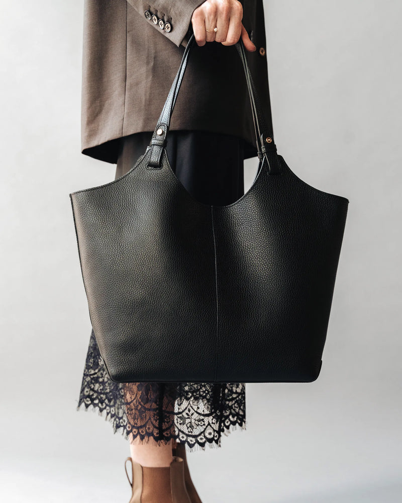 Woman in gray blazer and lace-hem dress holds a large pebbled leather tote. The Lisboa Tote Black.