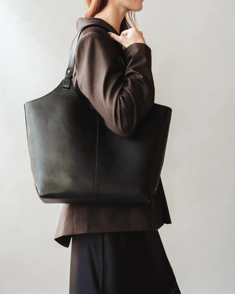 Woman in dark blazer carries a large pebbled leather tote on her shoulder. The Lisboa Tote Black.