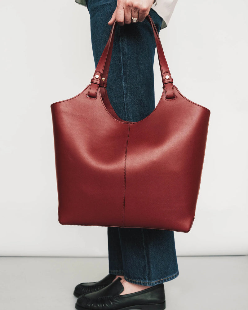 Leather tote bag held by handles, showcasing structured silhouette and gold hardware. The Lisboa Tote Resort Red.