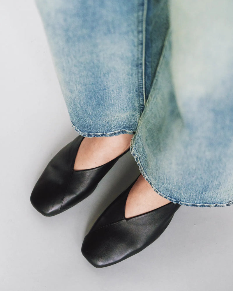 Leather square-toe flats on woman's feet with light blue jeans. The Marché Flat Black. Sizes 5–12, available in wide.
