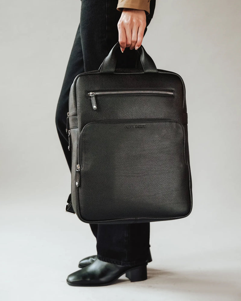 Woman pants holds pebbled leather structured backpack by its top handle. The Refined Backpack Black.