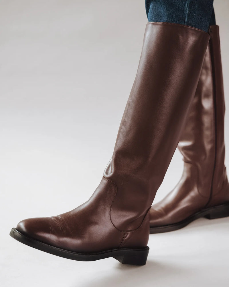 Woman in dark jeans wears espresso brown leather knee-high riding boots with flat heel. The Riding Boot Espresso. Sizes 5–12.