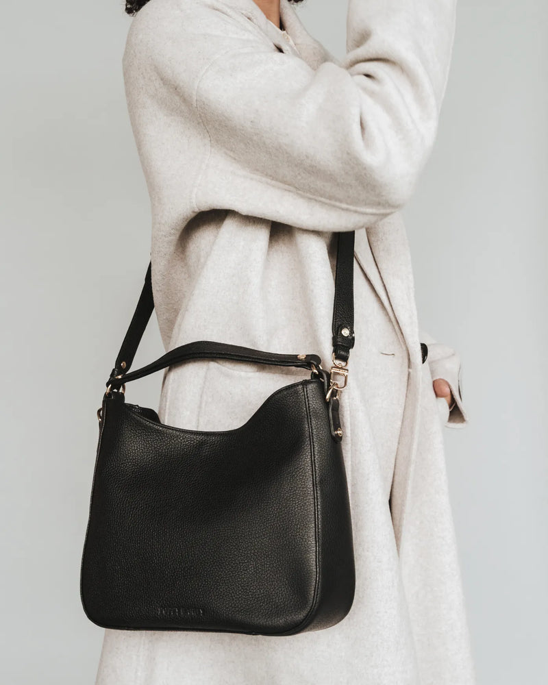 Woman in cream coat wears pebbled leather hobo bag with adjustable strap over shoulder. The Sixteenth Tote Black.