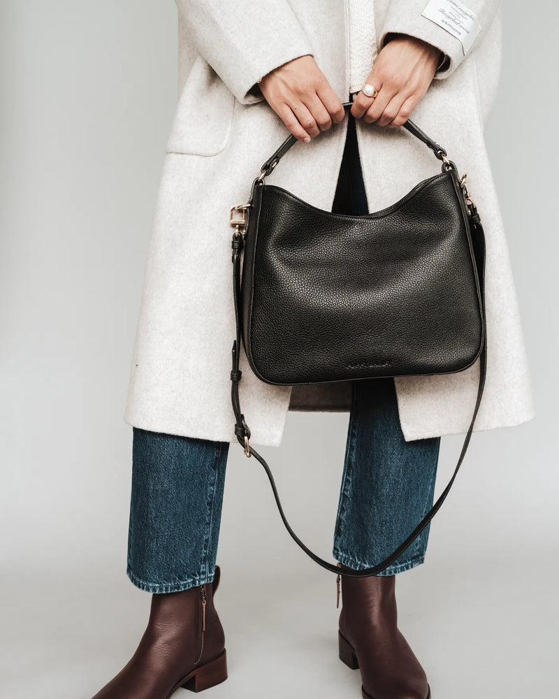 Woman in cream coat and jeans holds pebbled leather hobo bag by its handle. The Sixteenth Tote Black.