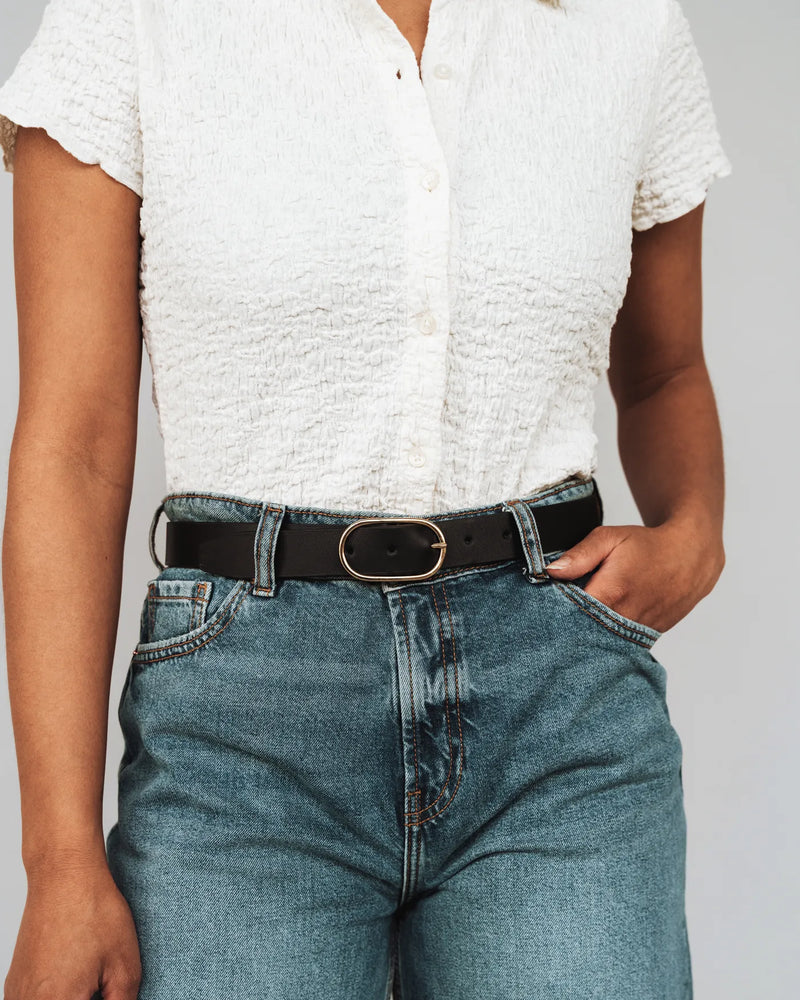 Woman in white blouse and jeans wears a black leather belt with oval gold buckle. The Sleek Belt Gold Black.
