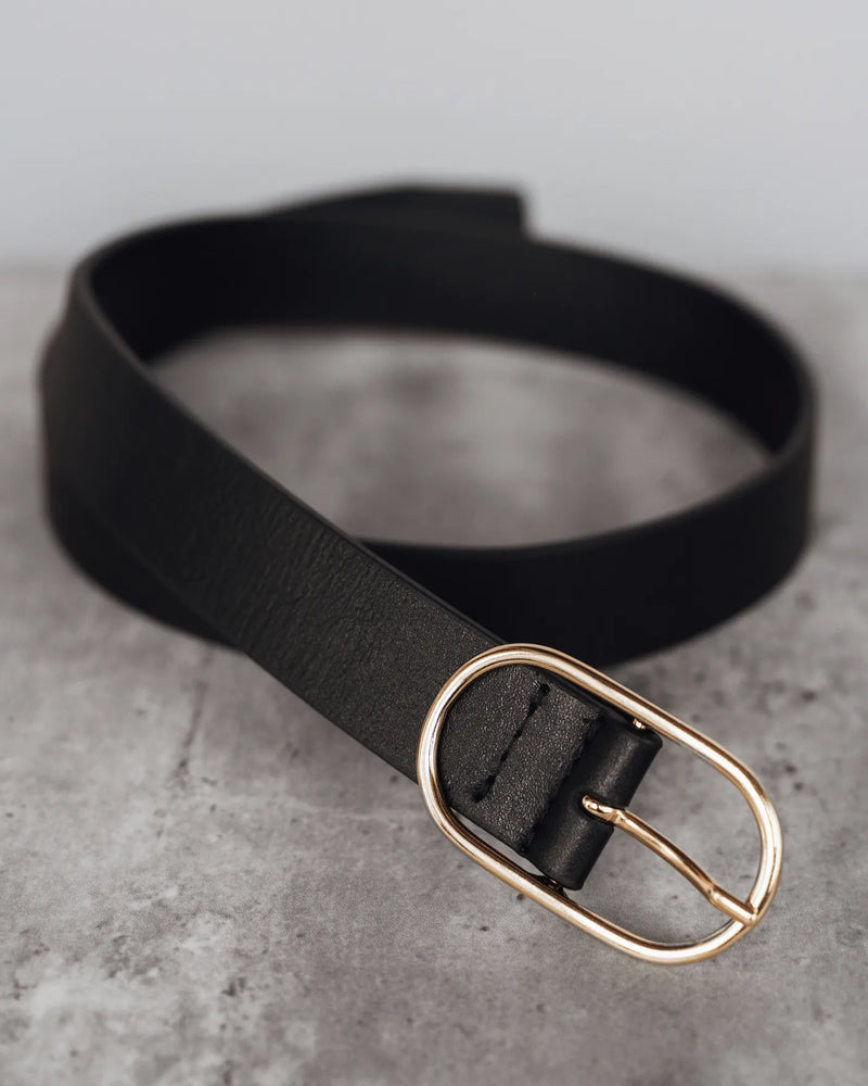 Black leather belt with oval gold-tone open buckle, coiled on a stone surface. The Sleek Belt Gold Black.