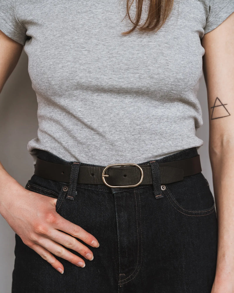 Woman in grey tee and dark jeans wears a black leather belt with oval gold buckle. The Sleek Belt Gold Black.