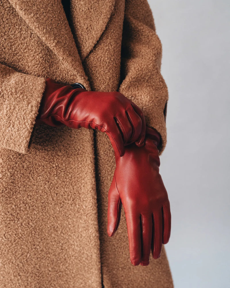 Woman in camel coat wears merlot red leather gloves with ruched wrist gathered detail. The Sleek Leather Glove Merlot.