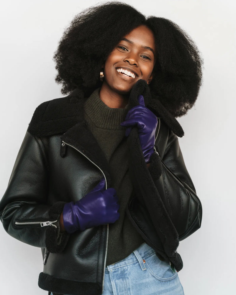 Woman smiling in black biker jacket, olive turtleneck, leather gloves. The Sleek Leather Glove Royal Blue.