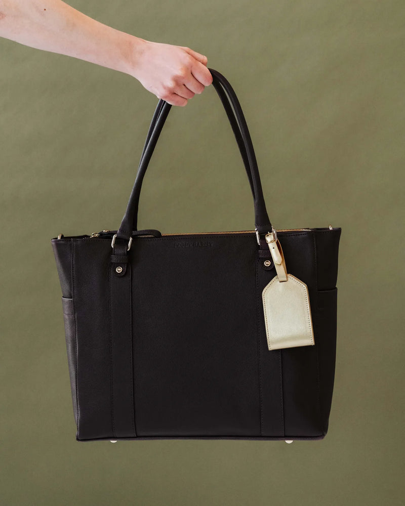 Metallic leather luggage tag on a black structured tote held by a woman. The Slim Luggage Tag Champagne.