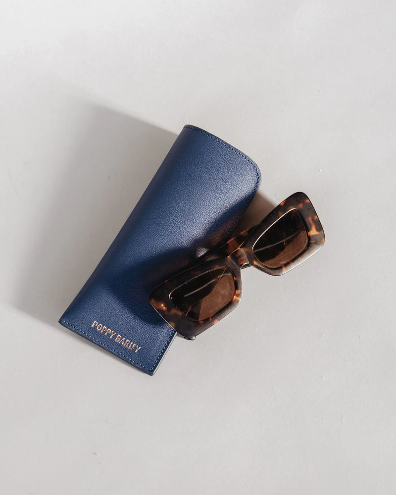 Navy leather sunglasses sleeve with gold foil branding beside tortoiseshell sunglasses. The Sunglasses Sleeve Pacific.