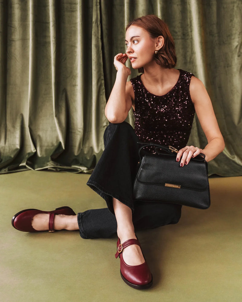 Woman seated wearing resort red Mary Jane flats with a sequin top and bag. The Today Mary Jane Resort Red. Sizes 5–12.