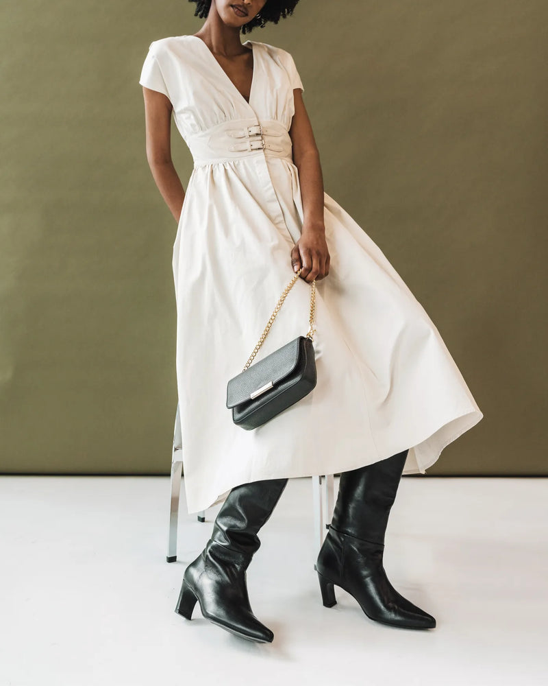 Woman in cream dress wearing leather tall boots and holding black chain bag. The Toujours Tall Boot Black. Sizes 5–12.